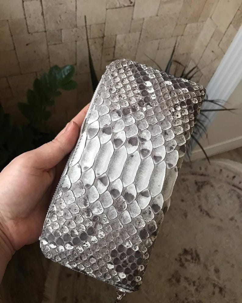 Op de afbeelding: Een rechthoekige portemonnee met een gestructureerd slangenleerpatroon in grijze, witte en zwarte tinten. De portemonnee heeft een ritssluiting rondom met een zilverkleurige metalen ritstrekker en een gedetailleerd schubbenpatroon.