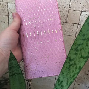 May include: A rectangular pink wallet with a textured snakeskin pattern and subtle gold foil accents. The wallet features a full-length zip closure along the side and top, with a metallic zip pull tab visible at the bottom corner.