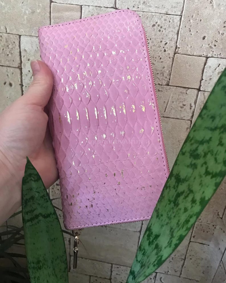 May include: A rectangular pink wallet with a textured snakeskin pattern and subtle gold foil accents. The wallet features a full-length zip closure along the side and top, with a metallic zip pull tab visible at the bottom corner.