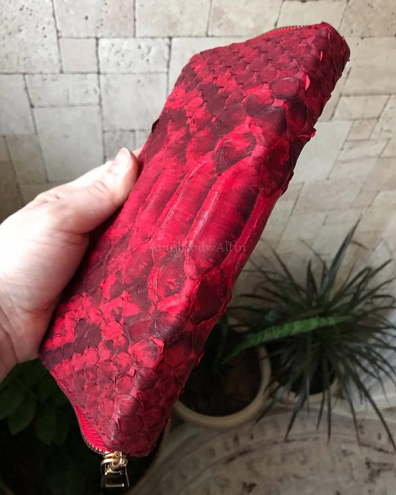 Peut inclure: Une pochette rectangulaire &agrave; fermeture &eacute;clair pr&eacute;sentant une texture en peau de serpent rouge vif et cramoisi fonc&eacute;. L'ext&eacute;rieur affiche un motif d'&eacute;cailles distinct avec une finition brillante, compl&eacute;t&eacute; par une tirette dor&eacute;e.