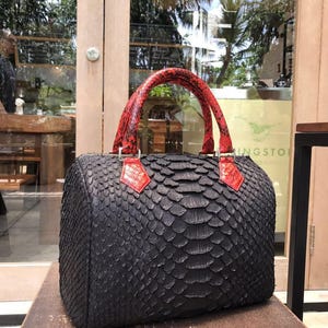Puede incluir: Un bolso estructurado con un exterior negro texturizado con un patrón de escamas repetitivo. El bolso incluye dos asas de color rojo vibrante con una textura de escamas a juego, unidas al cuerpo principal con pestañas triangulares rojas.