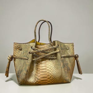 May include: A metallic gold handbag with a textured snakeskin pattern, featuring two top handles and a decorative drawstring cord that ties in knots on both sides. The interior lining is a bright yellow, and the bag has a structured base.