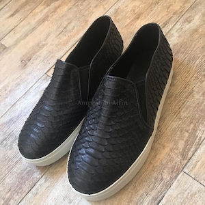 May include: A pair of black slip-on sneakers featuring a textured snakeskin-like pattern across the upper, elastic side gussets, and a thick white rubber sole. The word AmphibioAlfin is printed in small, faint letters on the side of the shoes.