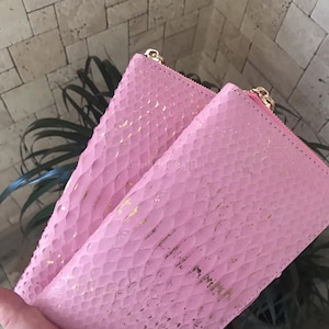 May include: A rectangular pink wallet featuring a textured snakeskin-like pattern with gold-colored foil accents. The wallet has a gold-colored metal zipper closure along the top edge and a smooth pink interior lining visible at the opening.
