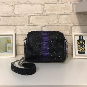 May include: A rectangular shoulder bag with a snakeskin texture, featuring a deep purple center stripe that fades into black on the sides. The bag has a silver chain strap, a black leather shoulder pad, and white paint splatters across the surface.