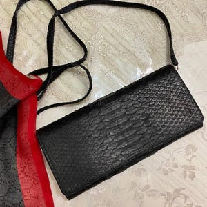 May include: A black rectangular crossbody bag with a textured snakeskin pattern and a thin shoulder strap. Beside the bag is a red and black scarf featuring a repeating logo pattern. The bag has a flap closure and a structured rectangular shape.