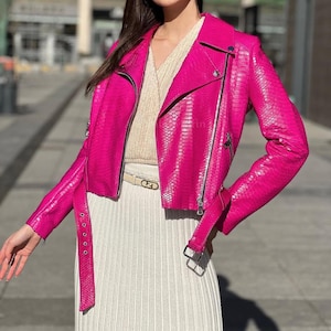 Peut inclure: Une veste motard rose vif avec une texture peau de serpent, dotée de fermetures éclair argentées, d'un revers cranté et d'une ceinture à boucle assortie à la taille. La veste est portée sur un haut en maille côtelée beige et une jupe assortie.