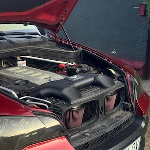 Könnte beinhalten: Ein rotes Fahrzeug mit geöffneter Motorhaube, das einen Hochleistungsmotorraum freigibt. Der Motor verfügt über ein schwarzes Lufteinlasssystem mit zwei markanten konischen roten Luftfiltern und einer schwarzen Abdeckung mit weißem Adler-Logo.