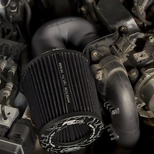 Pode incluir: Um filtro de ar cilíndrico preto com textura de malha e o texto Hyper Flow FMIC Pro AIR FILTER impresso na lateral. O filtro possui uma tampa circular com o texto FMIC Pro e TUNING PARTS, conectado a um tubo de admissão preto.
