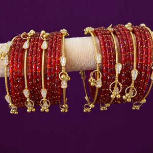 Peut inclure: Un ensemble de deux groupes de bracelets rigides en verre rouge à facettes avec des bandes en métal doré. Chaque bracelet comporte de petites breloques dorées avec des cristaux transparents en forme de goutte suspendus aux bords.