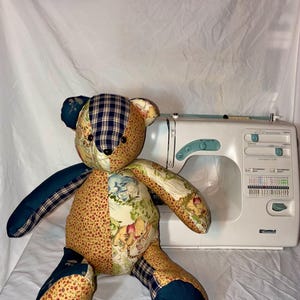 May include: A patchwork teddy bear constructed from various fabrics including blue, yellow, and floral patterns, featuring a Winnie the Pooh print on the torso, positioned in front of a white Kenmore sewing machine with teal control dials.