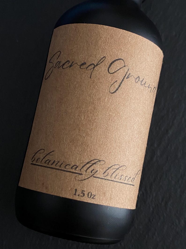 Pode incluir: Uma garrafa preta com um r&oacute;tulo castanho com as palavras "Sacred Ground" e "Botanically Blessed" em escrita elegante. O r&oacute;tulo tamb&eacute;m indica um volume de 44 ml. A garrafa tem um design elegante.