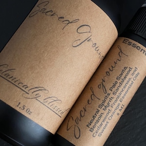 Pode incluir: Duas garrafas pretas de &oacute;leo essencial com r&oacute;tulos castanhos. Os r&oacute;tulos dizem "Sacred Ground" em it&aacute;lico. Uma garrafa tem 1,5 oz, a outra 8 ml. Ingredientes: Palo Santo, Benjoim e &oacute;leos essenciais de laranja doce.