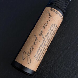 Pode incluir: Um frasco de spray de &oacute;leo essencial preto com um r&oacute;tulo castanho. O r&oacute;tulo diz "Sacred ground" em it&aacute;lico, com as palavras "Neutral Spirits, Palo Santo, Benzoin, Myrrh, Sandalwood, Cedarwood, Frankincense, and Sweet Orange Essential Oils."