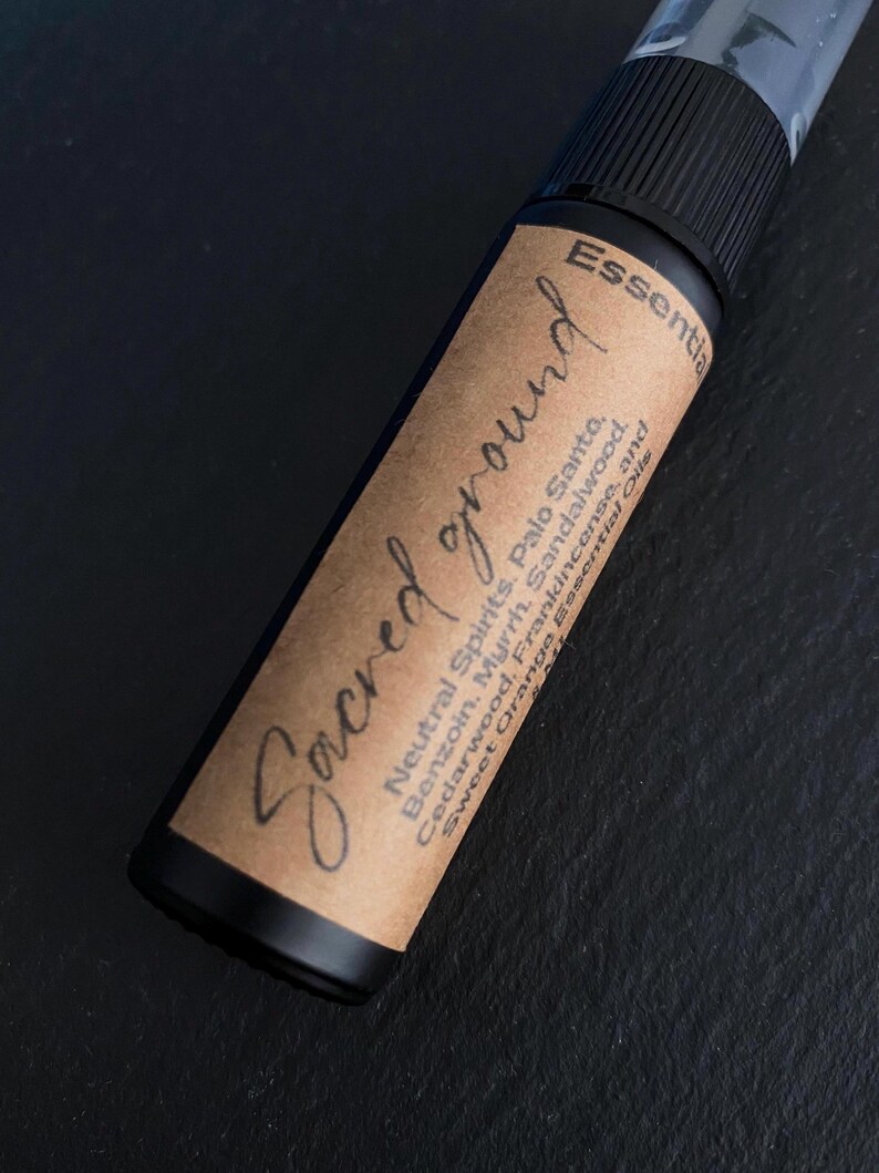 Pode incluir: Um frasco de spray de &oacute;leo essencial preto com um r&oacute;tulo castanho. O r&oacute;tulo diz "Sacred ground" em it&aacute;lico, com as palavras "Neutral Spirits, Palo Santo, Benzoin, Myrrh, Sandalwood, Cedarwood, Frankincense, and Sweet Orange Essential Oils."