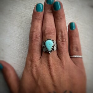 May include: A silver ring with a turquoise stone set in a teardrop shape. The ring is adorned with small silver beads and has a delicate design.
