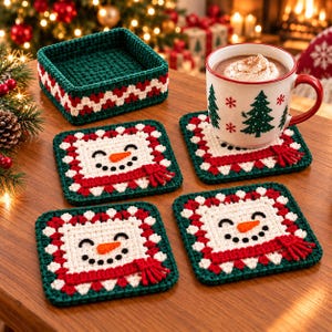 Puede incluir: Un juego de tres posavasos cuadrados de ganchillo con una cara de muñeco de nieve blanca, nariz de zanahoria y bufanda roja, bordeados por patrones de zigzag rojos y blancos sobre un borde verde oscuro, con una cesta cuadrada a juego.
