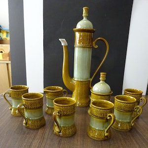 May include: A vintage ceramic coffee set in shades of brown and green. The set includes a tall coffee pot, a creamer, a sugar bowl, and six mugs. The coffee pot has a long spout and a curved handle. The mugs have handles and a textured design.