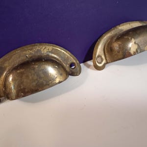 May include: Two antique brass drawer pulls with a half-moon design. The pulls have a tarnished, aged appearance with visible wear and tear, including areas where the brass finish has worn away. Each pull has two holes for mounting.