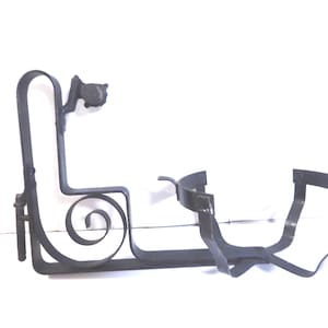 May include: Black wrought iron wall-mounted wine bottle holder. The holder features a decorative scroll design and a curved cradle to secure the bottle. The design includes a small, round knob and a bracket for mounting.