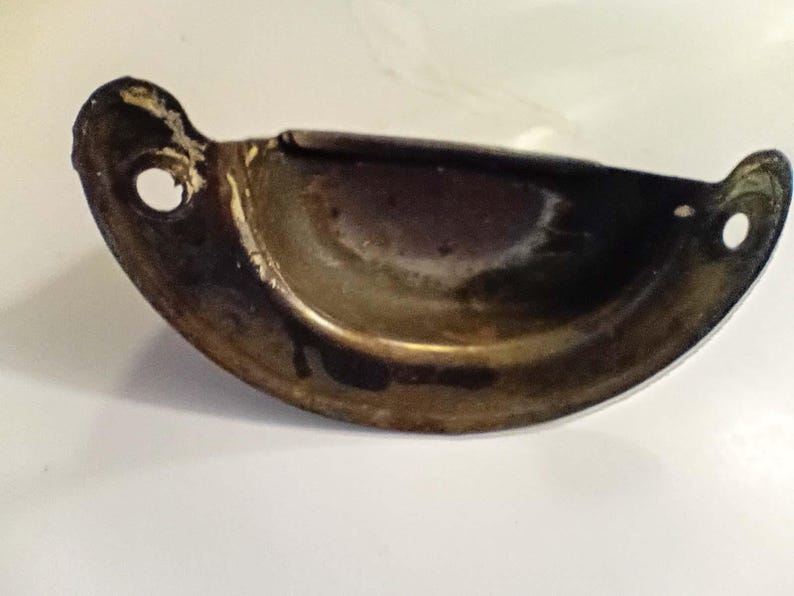 K&ouml;nnte beinhalten: Ein halbmondf&ouml;rmiger, vintage Schubladengriff mit dunkler, gealterter Messingoberfl&auml;che. Der Griff hat an beiden Enden zwei kleine L&ouml;cher zur Befestigung und einen gebogenen, vertieften Griffbereich. Das Metall weist Gebrauchsspuren und Oxidation auf.