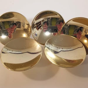 May include: Five gold-colored, reflective, circular objects are arranged in a cluster. The objects have a concave shape, with a polished, mirror-like surface. The reflections show a person holding a phone.