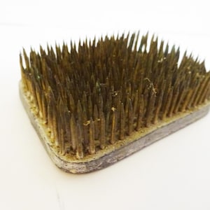 May include: A square, vintage flower frog with numerous sharp, bronze-colored pins. The pins are densely packed and protrude from a gray metal base. This floral arranging tool is designed to hold flower stems in place.