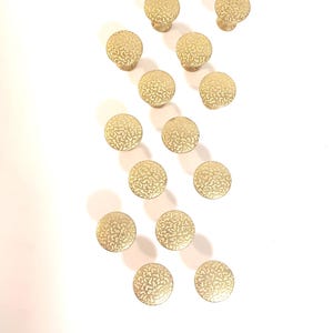 May include: A collection of twelve gold-colored cabinet knobs. Each knob has a round, textured face and a short stem. The knobs are arranged on a white surface, casting soft shadows. The knobs are likely for furniture.