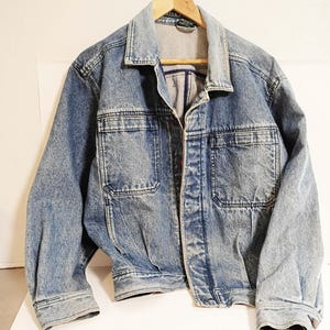May include: A light blue denim jacket with two chest pockets and two side pockets. The jacket is hanging on a wooden hanger. The denim has a faded, worn look. The jacket has a button closure and a collar.