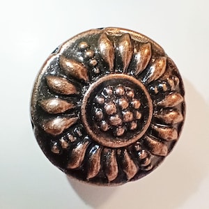 May include: A round metal cabinet knob featuring a raised, three-dimensional sunflower design. The surface has a dark, antiqued copper finish with highlights on the petals and central seeds, creating a textured, metallic appearance.