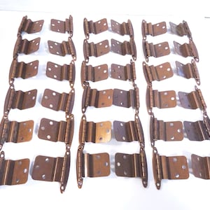 May include: A collection of antique bronze-colored cabinet hinges. The hinges are in various stages of being opened and closed, with multiple screw holes visible. The hardware is arranged on a white surface.