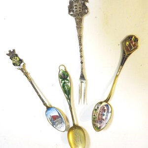 May include: Four antique spoons and a fork are displayed against a white background. The spoons feature detailed enamel artwork, including a building scene, floral designs, and a coat of arms. The fork has a decorative handle.