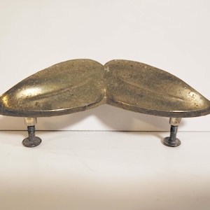 May include: A vintage brass-toned cabinet pull in the shape of a mustache. The pull has two curved, leaf-shaped sections connected in the middle. Each section is supported by a dark metal screw. The hardware is likely for a drawer or cabinet.