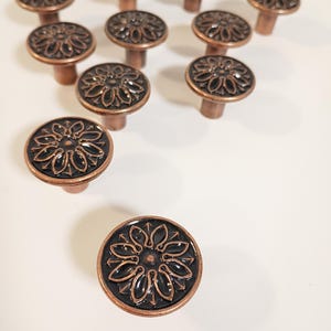 May include: A collection of bronze-colored cabinet knobs with a floral design. The knobs have a dark center and a raised, detailed pattern. The knobs are arranged on a white surface, showcasing their decorative appeal and potential for home decor projects.
