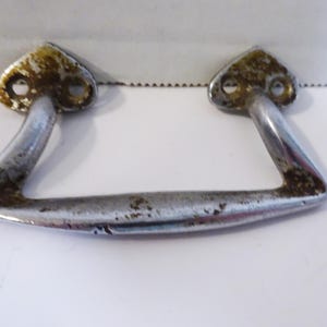 May include: A silver-toned metal handle with a curved shape and two holes on each side. The handle has a worn, aged look with some rust spots.