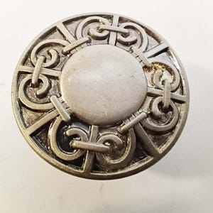 May include: A silver-toned, circular brooch with an intricate Celtic knot design. The brooch features a central, raised circle surrounded by interwoven patterns and geometric shapes. The metal has a slightly aged appearance.