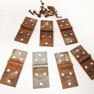 May include: An assortment of vintage metal hinges and screws. The hinges are rectangular with rounded edges, featuring a rusty brown patina. Some hinges are silver. The screws are small and bronze-colored, scattered above the hinges.