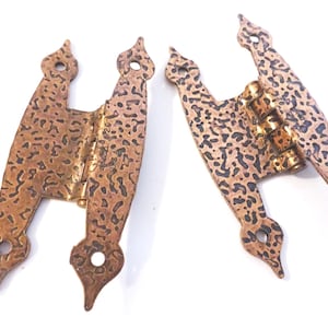 May include: Two antique copper-colored hinges with a textured surface and decorative finials. The hinges are in an open position, revealing the brass-colored hinge pins. Each hinge has three holes for mounting.