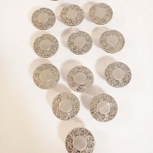 May include: A collection of silver-toned, circular buttons with an ornate, symmetrical design. Each button features a central, raised circle surrounded by a detailed, swirling pattern. The buttons are arranged on a white surface.