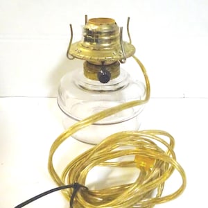 May include: A small, clear glass oil lamp with a brass-colored metal top and a black knob. The lamp has a coiled, gold-colored electrical cord. The lamp is designed to be used with oil.