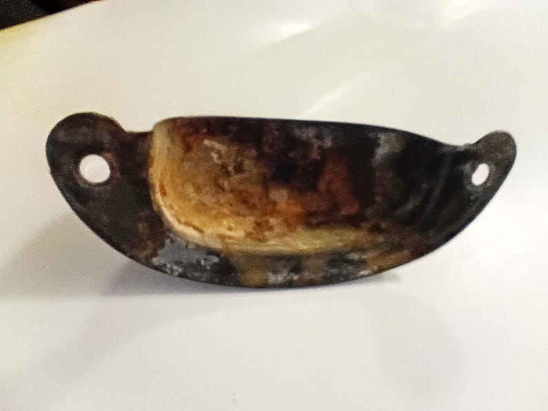 K&ouml;nnte beinhalten: Ein verwitterter Metall-Schubladengriff im antiken Stil mit geschwungenem, becherartigem Design. Der Griff hat ein dunkles, gealtertes Aussehen mit sichtbarem Rost und Abnutzung.