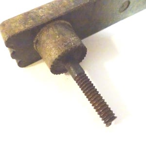 May include: Close-up of a weathered, antique metal hardware piece. The piece features a rectangular base with a cylindrical component and a threaded screw. The metal has a rusty, aged appearance, suggesting it's vintage.