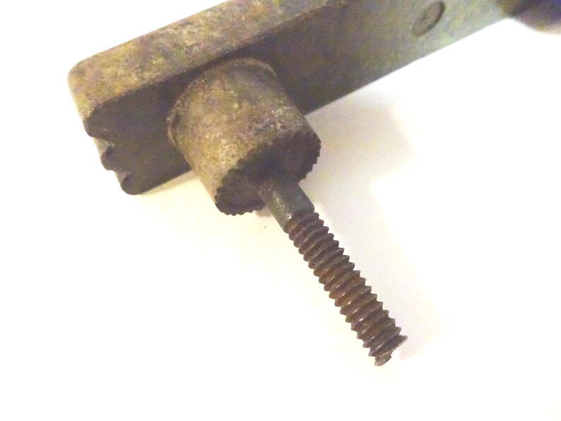 May include: Close-up of a weathered, antique metal hardware piece. The piece features a rectangular base with a cylindrical component and a threaded screw. The metal has a rusty, aged appearance, suggesting it's vintage.