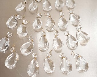 May include: A collection of clear, faceted crystal chandelier pendants. Each pendant has a teardrop shape with a smaller crystal bead and a gold-colored wire hook for hanging. The crystals are arranged on a reflective surface.