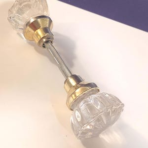 May include: Clear glass cabinet door knobs with a ribbed design. The knobs are attached to a metal rod with gold-colored metal accents. The knobs are likely for a cabinet or drawer.