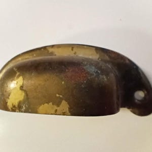 K&ouml;nnte beinhalten: Ein halbmondf&ouml;rmiger, antiker Schubladengriff mit Messing-Finish. Die Hardware hat eine dunkelbraune Patina mit abgenutzten gelben Farbflecken und einem einzelnen Befestigungsloch.