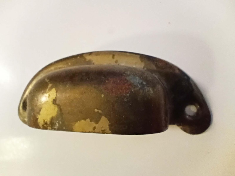 K&ouml;nnte beinhalten: Ein halbmondf&ouml;rmiger, antiker Schubladengriff mit Messing-Finish. Die Hardware hat eine dunkelbraune Patina mit abgenutzten gelben Farbflecken und einem einzelnen Befestigungsloch.