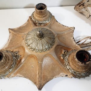 May include: A three-light ceiling fixture with a weathered, distressed beige and metallic finish. The central hub features a raised floral medallion, while each light socket is surrounded by a decorative laurel wreath motif and a chain.
