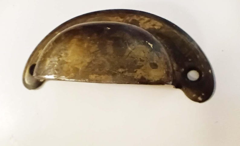 K&ouml;nnte beinhalten: Ein Vintage-Schubladengriff in Halbmondform mit einem angelaufenen, gealterten Messing-Finish. Die Hardware hat ein abgerundetes, becherartiges Design mit zwei Befestigungsl&ouml;chern und einem leicht gebogenen Griff. Abmessungen: ca. 10 cm breit.