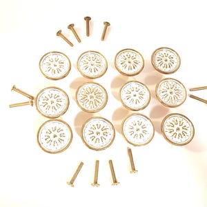 May include: A collection of decorative cabinet knobs and screws. The knobs are round with a white center and a gold-colored rim and design. The screws are gold-colored. The knobs and screws are arranged on a white surface.